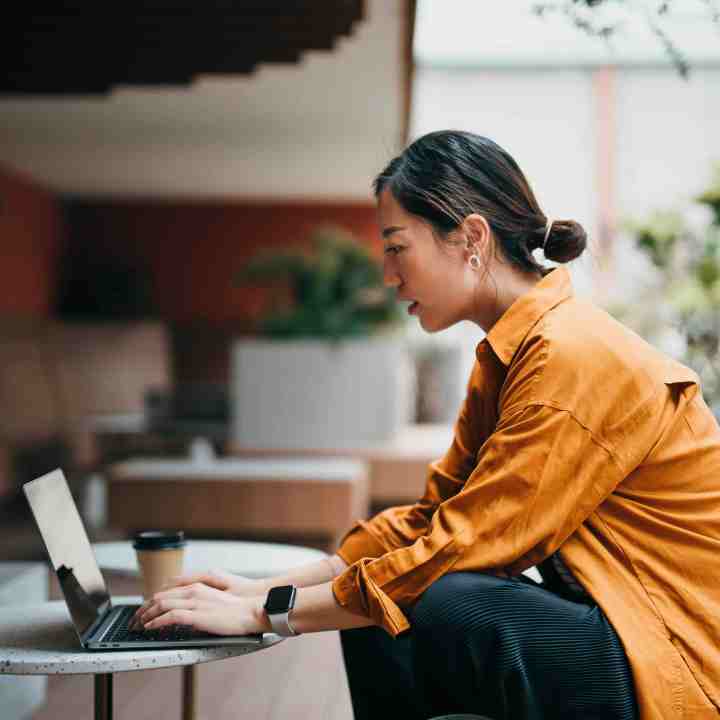 A woman in an orange shirt works on a laptop at an outdoor table.